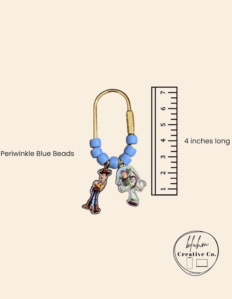 May include: A gold-toned metal keychain featuring a U-shaped clasp adorned with periwinkle blue beads and two character charms. A ruler next to the item indicates it is 4 inches long. The text Periwinkle Blue Beads is visible near the beads.