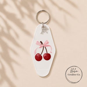 May include: White motel-style keychain with a cherry design. The keychain features two red cherries with a pink bow and a silver key ring. The bottom right corner has a circular logo with the text "Bluhm Creative Co."