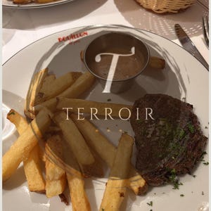 May include: A plate of food featuring a steak, french fries, and a small metal cup of sauce. The plate has the word "TERROIR" printed on it. A basket of bread is in the background.