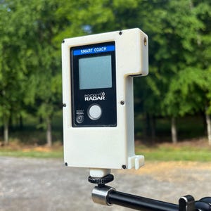 May include: A white "Smart Coach" device with a black screen and the "Pocket Radar" logo, mounted on a black adjustable arm. The device is designed for sports training, with a button labeled "Recall". The background features green trees and a blue sky.
