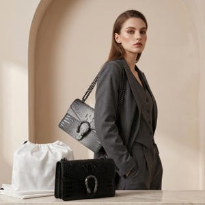 May include: Two leather handbags with a crocodile texture and a metal horseshoe-shaped clasp. One bag is black, the other is grey. A woman in a grey suit stands behind the bags. A white drawstring bag is on the table.