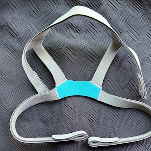 May include: A gray and teal CPAP headgear with adjustable straps and a teal center piece. The headgear is designed to fit around the head and under the nose, with a curved lower section.