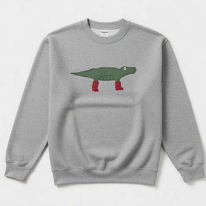 Puede incluir: Una sudadera gris jaspeada con cuello redondo, con un cocodrilo verde de dibujos animados con botas rojas. El cocodrilo es el elemento central del diseño en la parte delantera de la parte superior de manga larga. Diseño sencillo e informal.