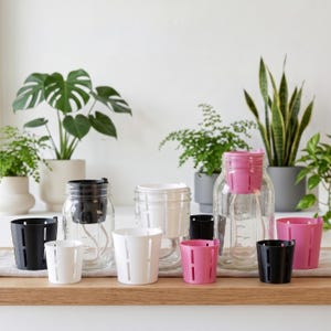 May include: A collection of self-watering planters consisting of clear glass jars and removable plastic inserts in black, white, and pink. Each insert features vertical slits and a white wick for water absorption, designed for indoor plant care.
