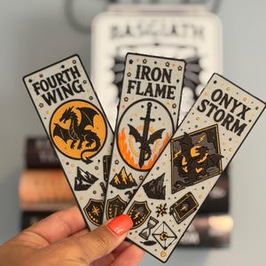 May include: Three rectangular bookmarks featuring black and orange illustrations. The first reads Fourth Wing with a dragon, the second Iron Flame with a sword and fire, and the third Onyx Storm with a dragon and lightning. Each has small star accents.
