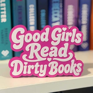May include: A bright pink, three-dimensional sign with white lettering that reads Good Girls Read Dirty Books. The sign features a bold, rounded font style with a small heart icon centered between the second and third lines of text.