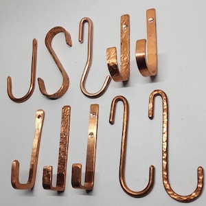 May include: A collection of ten copper hooks in various shapes, including S-hooks and J-hooks. Some hooks feature a smooth, polished finish, while others display a textured, hammered surface. Each hook includes pre-drilled holes for mounting.