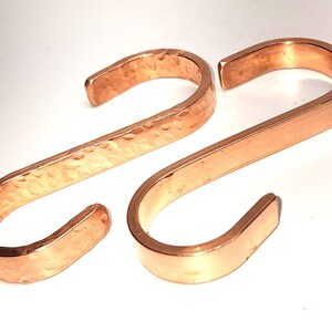May include: Two S-shaped hooks made of polished copper. One hook features a textured, hammered finish across its surface, while the other hook has a smooth, plain finish. Both hooks are curved at each end to form a distinct S-shape design.