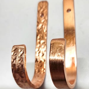 May include: Two J-shaped wall hooks made of polished copper. The hook on the left features a textured, hammered surface finish, while the hook on the right has a smooth, brushed metal finish. Each hook includes a single circular mounting hole at the top.