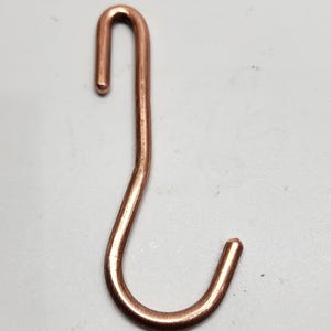 May include: A metallic copper-colored S-shaped hook with a smooth, polished finish. The hook features a curved top section and a wider curved bottom section, both with rounded ends, designed for hanging items securely on a rod or rail.