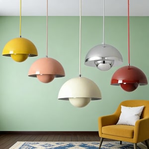 May include: A collection of five modern pendant lights in various colors, including yellow, peach, cream, silver, and red. Each light has a rounded shade and a spherical bottom, suspended from the ceiling by a cord. A yellow armchair with a white pillow is in the foreground.