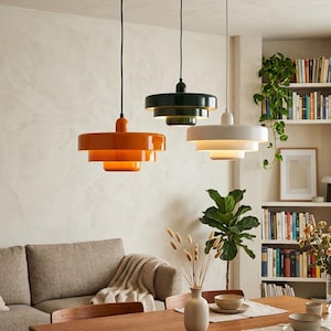 Può includere: Tre moderne lampade a sospensione arancioni, verde scuro e bianche pendono dal soffitto. Ogni lampada ha un design circolare a più livelli. Le luci sono sospese sopra un tavolo da pranzo in legno, con un divano e una libreria sullo sfondo.