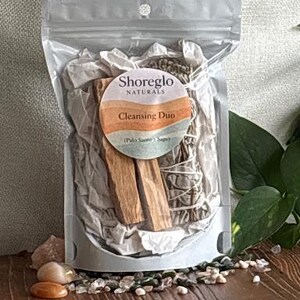May include: A clear plastic bag containing a cleansing duo kit. The kit includes two pieces of Palo Santo wood, a sage bundle, and a round label that reads "Shoreglo Naturals Cleansing Duo (Palo Santo / Sage)". The kit is packaged with white tissue paper.