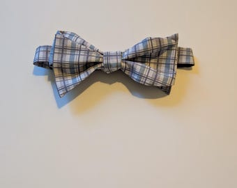 May include: A pre-tied bow tie featuring a plaid pattern in shades of light blue, white, and gray. The fabric is folded into a structured, symmetrical shape with a central knot and an adjustable strap visible on the sides.