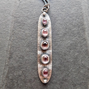 May include: A long, silver pendant necklace with five oval, pink gemstones set in individual bezels. The pendant has a hammered texture and a small loop at the top for the chain. The chain is a dark, oxidized silver.