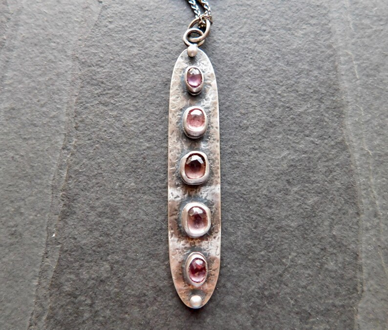 May include: A long, silver pendant necklace with five oval, pink gemstones set in individual bezels. The pendant has a hammered texture and a small loop at the top for the chain. The chain is a dark, oxidized silver.