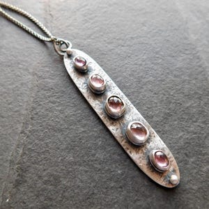 May include: A silver pendant necklace with a textured, elongated oval pendant. The pendant features five oval, pink gemstones. The necklace chain is a delicate silver. The pendant is approximately 3 inches long.