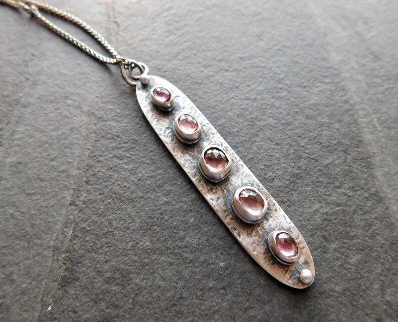 May include: A silver pendant necklace with a textured, elongated oval pendant. The pendant features five oval, pink gemstones. The necklace chain is a delicate silver. The pendant is approximately 3 inches long.