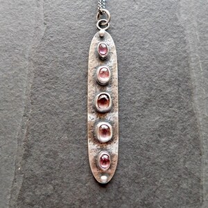 May include: A long, silver-toned pendant necklace with five oval, pink gemstones. The pendant has a textured surface and a small loop at the top for the chain. The gemstones are set in individual bezels along the length of the pendant.