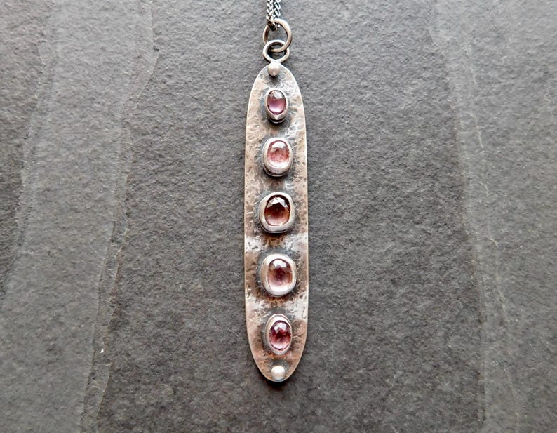 May include: A long, silver-toned pendant necklace with five oval, pink gemstones. The pendant has a textured surface and a small loop at the top for the chain. The gemstones are set in individual bezels along the length of the pendant.