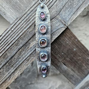 May include: A silver pendant necklace with five oval-shaped, pink gemstones set in a textured, elongated metal pendant. The pendant hangs from a delicate silver chain. The gemstones vary slightly in color, adding depth to the design.