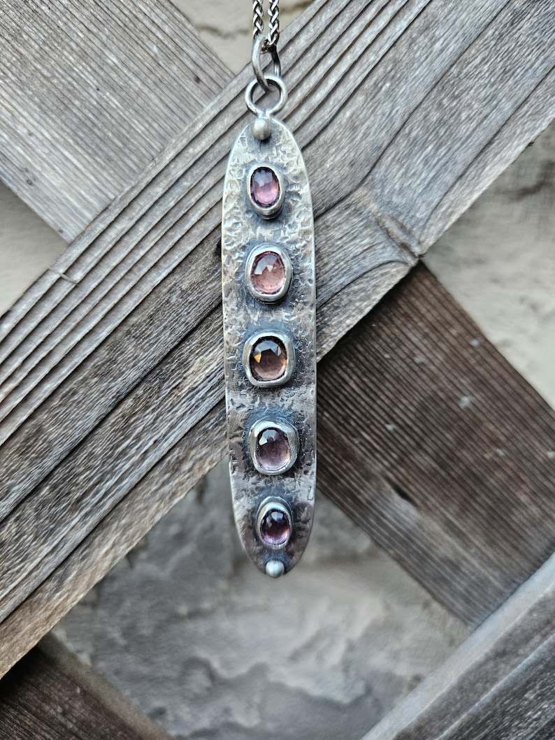 May include: A silver pendant necklace with five oval-shaped, pink gemstones set in a textured, elongated metal pendant. The pendant hangs from a delicate silver chain. The gemstones vary slightly in color, adding depth to the design.