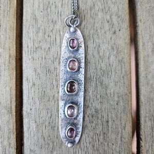 May include: A silver pendant necklace with a hammered texture. The pendant is an elongated oval shape, featuring five oval-shaped, light pink gemstones. The necklace chain is a delicate silver color, and the background is a wooden surface.