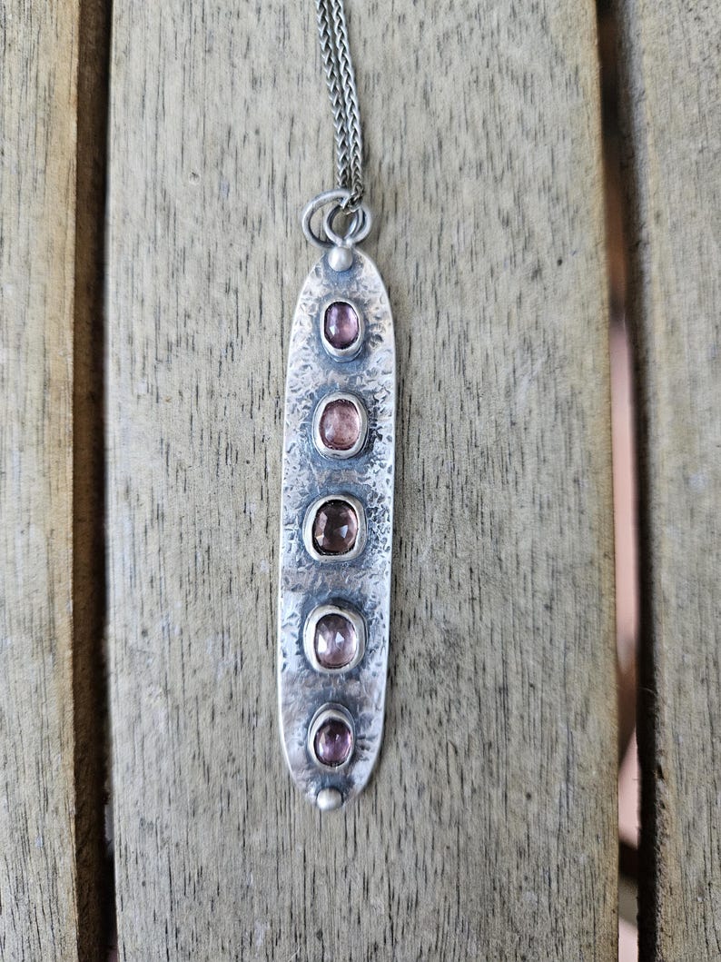 May include: A silver pendant necklace with a hammered texture. The pendant is an elongated oval shape, featuring five oval-shaped, light pink gemstones. The necklace chain is a delicate silver color, and the background is a wooden surface.