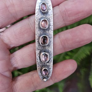 May include: A silver pendant necklace with five oval-shaped, pink gemstones set in a textured, elongated silver pendant. The pendant hangs from a silver chain. The background is a blurred view of green foliage.