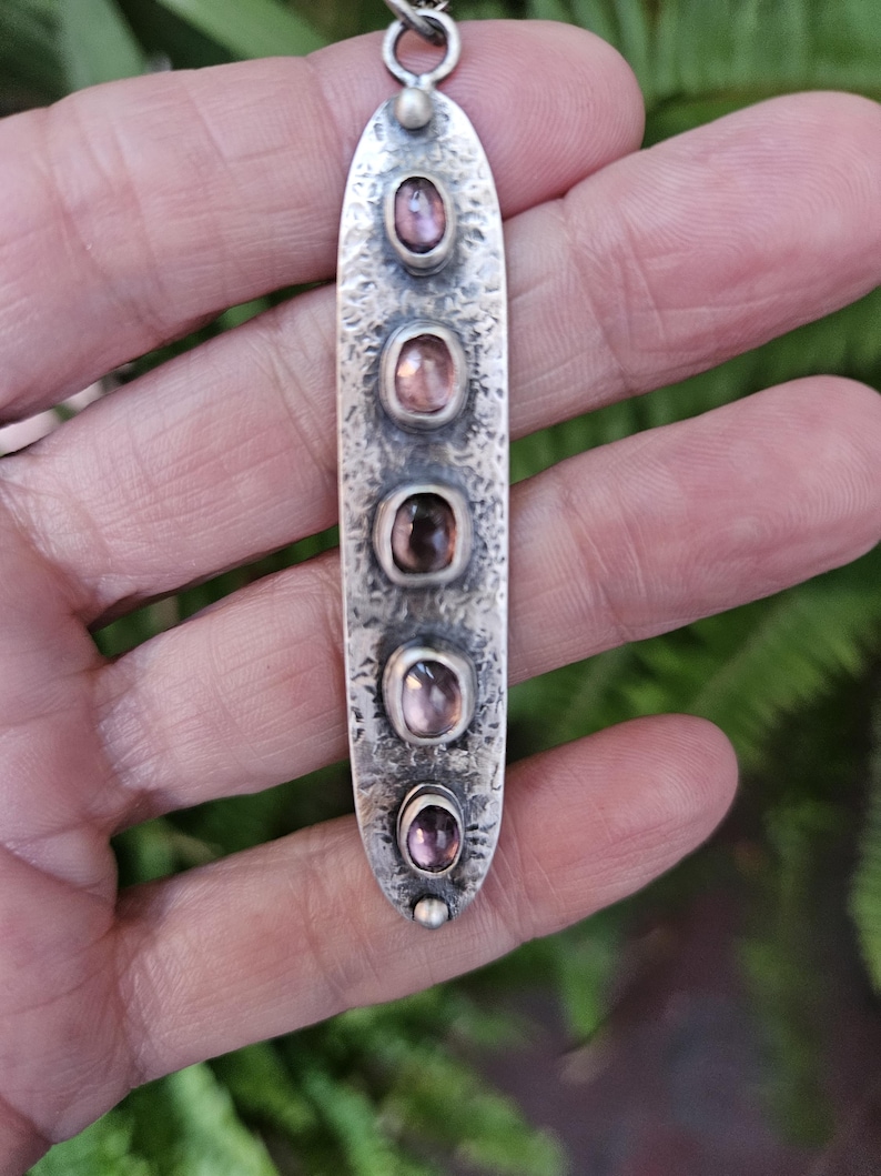 May include: A silver pendant necklace with five oval-shaped, pink gemstones set in a textured, elongated silver pendant. The pendant hangs from a silver chain. The background is a blurred view of green foliage.