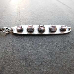 May include: A silver necklace featuring a horizontal bar pendant with five small, round, pink gemstones. The pendant is connected to a delicate chain. The necklace is displayed against a dark, textured background.