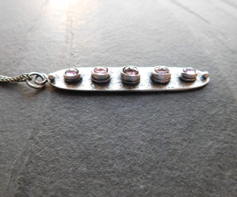 May include: A silver necklace featuring a horizontal bar pendant with five small, round, pink gemstones. The pendant is connected to a delicate chain. The necklace is displayed against a dark, textured background.