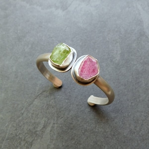 May include: A silver cuff bracelet with two raw gemstones set in silver. The gemstones are a light green and a pink.