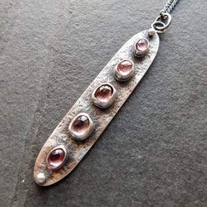 May include: A silver pendant necklace with a hammered texture. The pendant is an elongated oval shape and features five oval-shaped, pink gemstones. The necklace has a delicate chain and a small pearl at the bottom of the pendant.