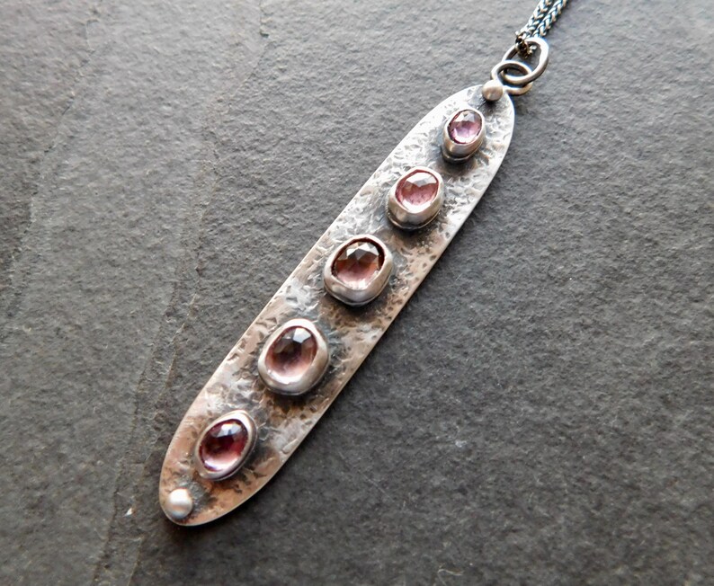 May include: A silver pendant necklace with a hammered texture. The pendant is an elongated oval shape and features five oval-shaped, pink gemstones. The necklace has a delicate chain and a small pearl at the bottom of the pendant.