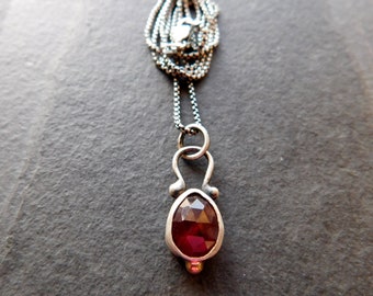 May include: A silver chain necklace with a pendant featuring a red gemstone set in a silver bezel. The pendant is shaped like a teardrop and has a small silver bead at the bottom.