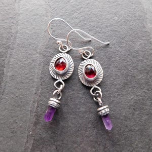 May include: Silver earrings with a red gemstone set in a silver circle and a purple amethyst pendant.
