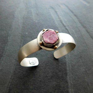 May include: A silver cuff bracelet with a pink gemstone set in a gold bezel. The bracelet has a hammered finish and is stamped with the number "14".