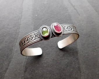 May include: A silver bracelet with an ornate design and two gemstones. The larger gemstone is pink and the smaller gemstone is green.