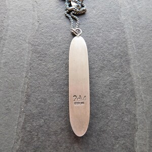 May include: A silver-toned pendant necklace with a long, slender, oval-shaped pendant. The pendant has the text "2di4 STERLING" engraved near the bottom. The necklace chain is a dark, textured metal.
