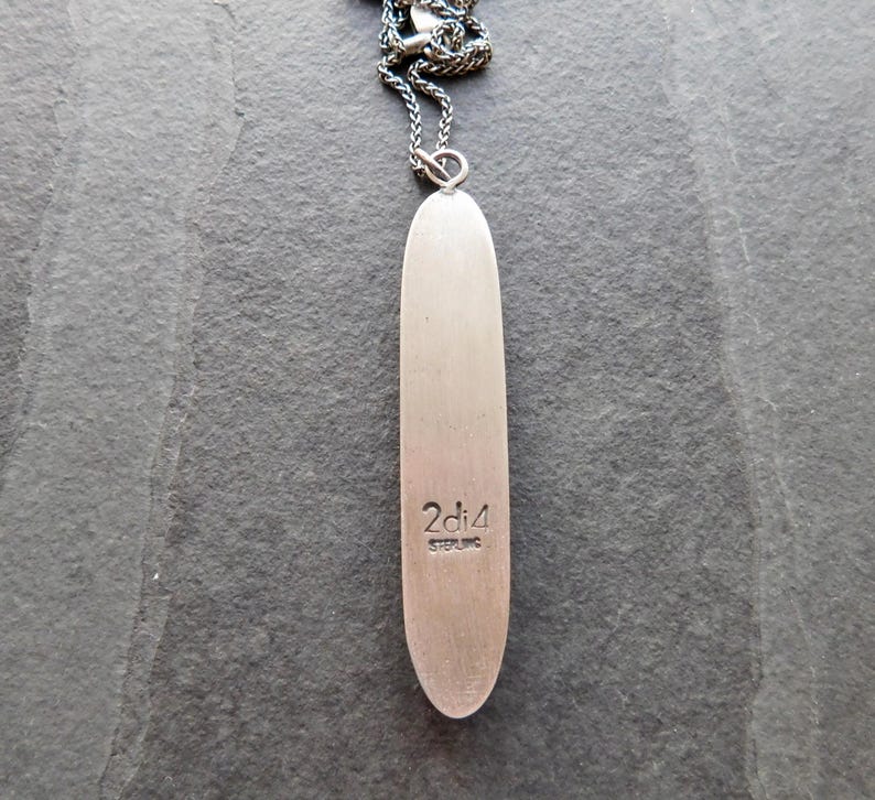 May include: A silver-toned pendant necklace with a long, slender, oval-shaped pendant. The pendant has the text "2di4 STERLING" engraved near the bottom. The necklace chain is a dark, textured metal.