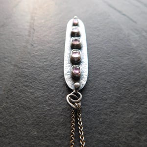 May include: A silver pendant necklace with five oval, pink gemstones set in a vertical row. The pendant has a textured surface and is attached to a delicate chain. The background is a dark, textured surface.