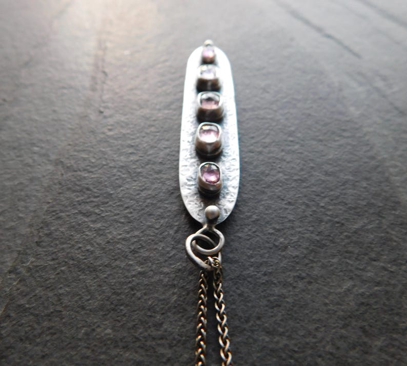 May include: A silver pendant necklace with five oval, pink gemstones set in a vertical row. The pendant has a textured surface and is attached to a delicate chain. The background is a dark, textured surface.