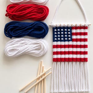 May include: A macrame wall hanging designed as an American flag with red, white, and blue cord, suspended from a wooden dowel. Included are four coiled bundles of cord in white, red, blue, and white, along with five additional wooden dowels.