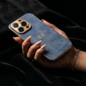 May include: A blue phone case with gold trim and a gold camera module is held in a hand. The case has the initials "B.K." in gold lettering. The background is a dark, velvety fabric.