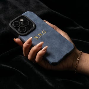 Op de afbeelding: Een blauwe telefoonhoes met gouden initialen "S.N.G" wordt in een hand gehouden. De hoes heeft een getextureerd uiterlijk en een zwarte cameralensbeschermer. Een gouden armband is zichtbaar om de pols. De achtergrond is een donkere, fluwelen stof.