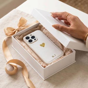 May include: A white phone case with a gold heart design inside a white gift box. The case has the initials "M.J" in gold. The box is lined with brown paper and tied with a gold ribbon.