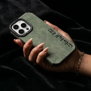 May include: An olive green phone case with a black camera ring and black trim. The case has a textured surface and features black Arabic text. The phone case is held in a hand with a gold bracelet.