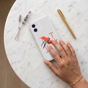 Puede incluir: Funda de teléfono transparente con un diseño de letra "N" roja y el nombre "Maar" en escritura negra. La funda está sobre una superficie de mármol blanco con un bolígrafo dorado y ramitas de lavanda.