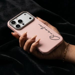 May include: A pale pink phone case with the word "Queenation" in black script. The phone case has a silver camera border. The phone is held in a hand with a gold bracelet.